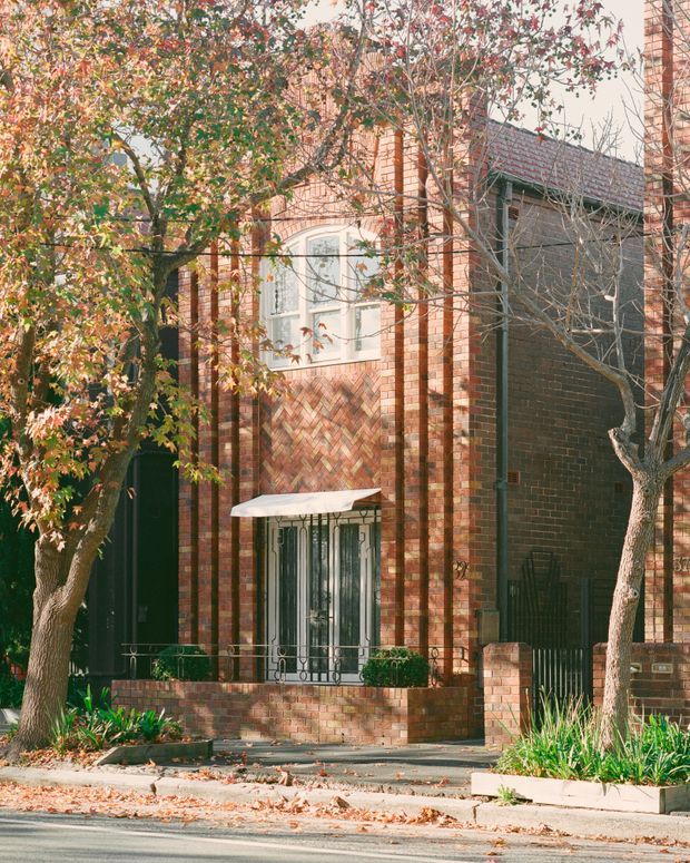 Street-facing view of a traditional brick townhouse facade with strong vertical brick piers, decorative herringbone brickwork, arched and divided window detailing, a dark recessed entry, wrought-iron security bars, and a small raised front garden behind a low brick wall. Mature deciduous trees and planted garden beds frame the frontage.