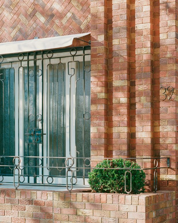 Close-up exterior view of a brick residential facade featuring a recessed entry door with textured glass, decorative metal security grille, brick planter ledge, and a neatly trimmed shrub. The brickwork includes patterned herringbone detailing and projecting vertical pilasters, with a visible house number.