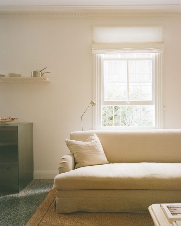 A softly lit, refined living room with a cream upholstered sofa positioned beneath a tall sash window with a Roman blind. The room features warm off-white walls, traditional decorative cornice and trim, a slim floor lamp, floating shelf with ceramics, dark green built-in cabinetry, and layered natural-texture rugs.