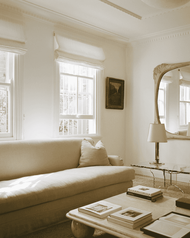 A bright, refined living room with cream upholstery, tall sash windows, ornate architectural mouldings, a large antique-style wall mirror, table lamp, glass console, and a low light-wood coffee table with books. The room has a warm, monochromatic palette and abundant natural light.