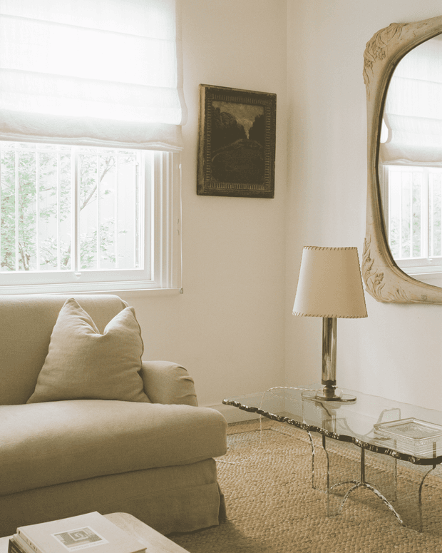 A softly lit, refined living room corner featuring a neutral upholstered armchair, glass coffee table with a sculptural metal frame, table lamp, textured natural-fibre rug, framed artwork, and an ornate wall mirror. Large windows with white Roman blinds bring in abundant daylight, creating a calm, warm and elegant atmosphere.