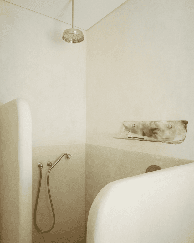 A softly lit minimalist bathroom featuring a curved freestanding bathtub in the foreground, a handheld shower fixture, a round rainfall showerhead, and a two-tone light cream and beige wall finish. A small metallic wall shelf or ledge adds a subtle aged accent.