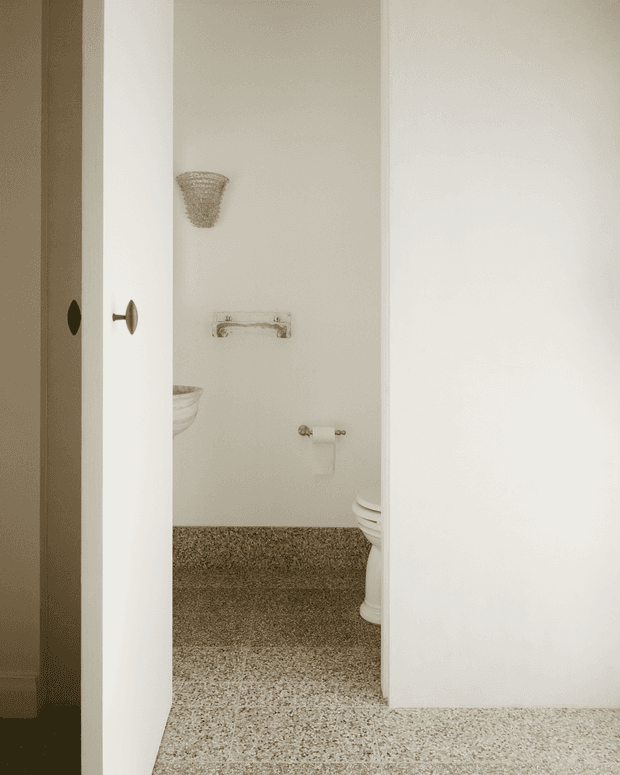 A narrow view into a compact, light-toned bathroom through a partially open white door. The room features cream walls, a textured beige carpeted floor, white ceramic sanitaryware, and aged brass or bronze fixtures including a towel rail and toilet-roll holder. A wall-mounted woven or textured light fixture adds a subtle decorative detail.