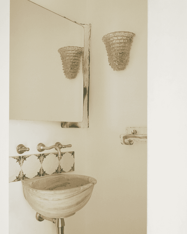 A close-up view of a compact, traditionally styled bathroom featuring a small sculptural stone or ceramic basin, exposed plumbing, aged metallic wall-mounted taps, a decorative patterned tile splashback, an ornate wall sconce, and a mirrored cabinet. The palette is warm off-white, beige, and antique brass tones.