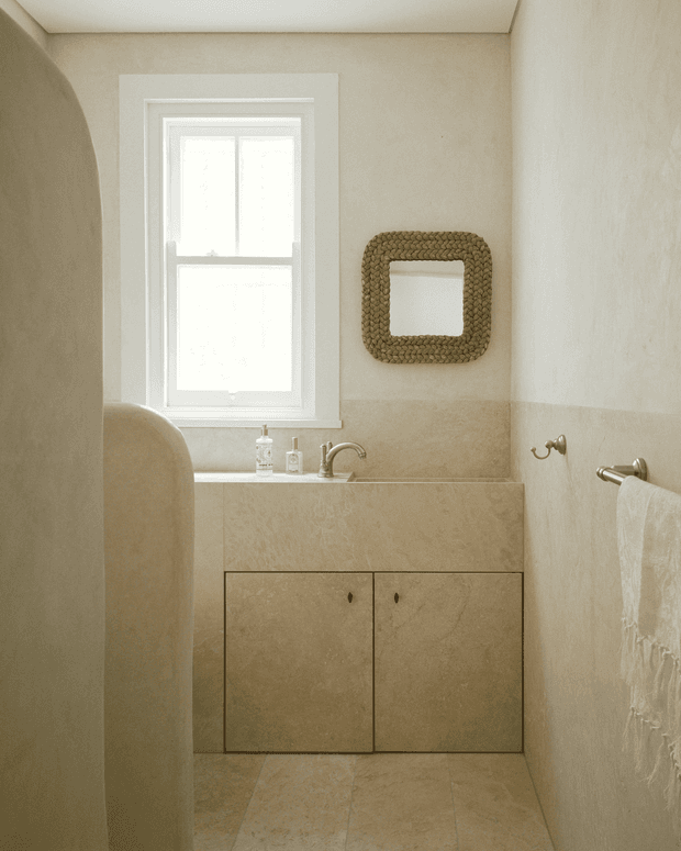 A compact, warm-toned bathroom with a white-framed window, integrated stone or plaster-look vanity, undermount basin and wall-mounted brushed metal tapware. The vanity has minimalist flat-front cabinetry, while a textured woven rectangular mirror frame and white towel add subtle detail. Beige tiled flooring and softly textured walls create a calm, monochromatic interior.