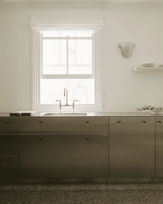 A softly lit traditional kitchen with a centered sash window above a sink, pale walls, ornate white window trim, dark muted cabinetry with simple knobs, and a textured carpeted floor. A small wall sconce and open shelf add understated decorative detail.