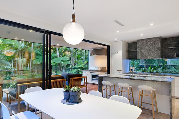 A bright, contemporary open-plan kitchen and dining area with a large white dining table in the foreground, a stone island with bar stools, integrated cabinetry, stainless-steel appliances, and dark-framed glazed doors opening to lush greenery. A large spherical pendant light hangs above the dining zone.