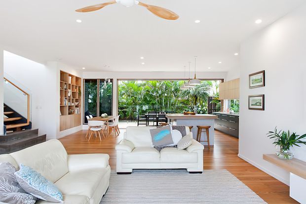 Bright open-plan contemporary living area with white sofas, a large pale textured rug, timber flooring, built-in timber shelving, and a compact dining area. The kitchen sits at the rear with a stone island and dark cabinetry, while full-height glazing opens toward lush greenery and an outdoor dining space. A timber staircase, recessed lighting, pendant lights, and a timber-bladed ceiling fan complete the interior.