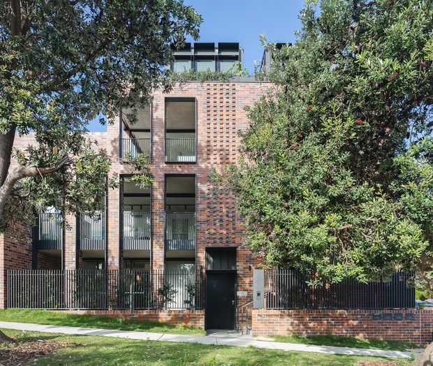 Street-facing view of a contemporary multi-storey residential building, partially screened by mature trees. The facade combines extensive red and charcoal brickwork with dark metal fencing, tall recessed windows, narrow balconies, and a rooftop terrace or balcony structure. A pedestrian entry gate and landscaped verge are visible at street level.