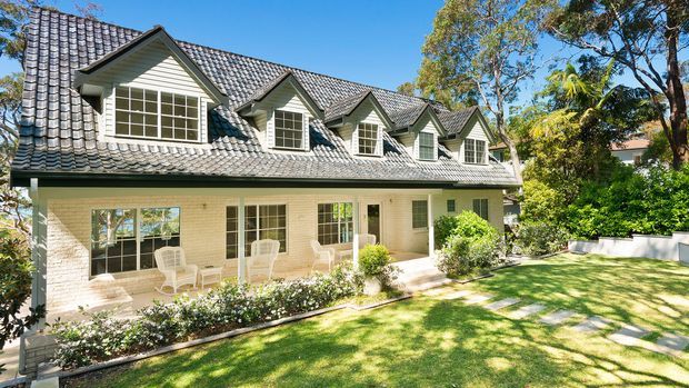 Exterior view of a large two-storey light-brick residence with a grey tiled pitched roof, repeated front-facing dormer windows, white-framed glazing, and a covered ground-floor verandah. The house opens onto a manicured lawn with established trees and shrubs in a suburban garden setting.