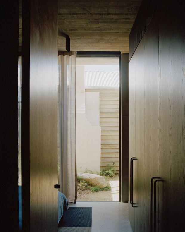 A narrow, dimly lit interior passage or entry hall lined with tall, dark timber built-in cabinetry and long black handles. A sheer curtain and open doorway lead to a small planted courtyard, while a partial blue upholstered seat and dark floor mat are visible in the foreground. The ceiling is clad in timber boards, creating a warm, intimate and understated atmosphere.