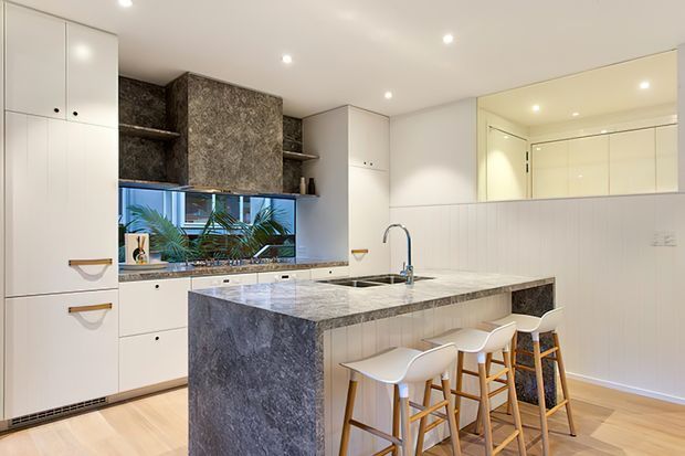 A bright contemporary kitchen with a large stone-clad island and integrated breakfast bar. White flat-panel cabinetry extends to the ceiling, complemented by a dark patterned stone backsplash and matching island sides. Three white bar stools with timber legs sit at the island, while a large mirror reflects the cabinetry and recessed ceiling lighting. Pale timber flooring and abundant integrated downlights create a clean, warm, open feel.