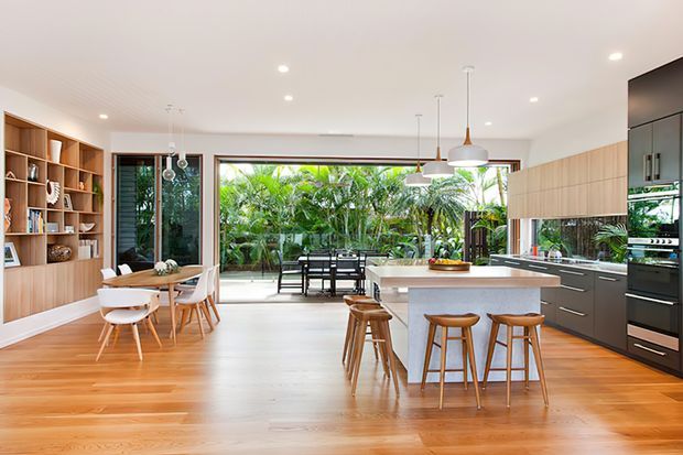 Bright open-plan contemporary kitchen and dining area with warm timber flooring, a large pale island with timber bar stools, streamlined dark and timber cabinetry, integrated appliances, pendant lighting, and a built-in timber display wall. Full-height glazing opens to a lush tropical garden and outdoor dining area.