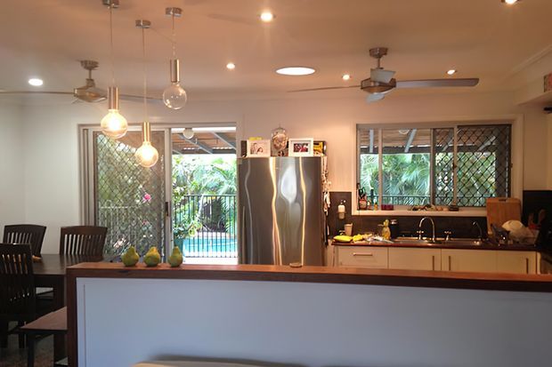 A warm, lived-in kitchen and dining area viewed across a large island or breakfast bar. The room has pale walls and cabinetry, dark stone-like benchtops, stainless-steel appliances, pendant lights, ceiling fans, tiled or smooth light flooring, and extensive windows and glazed doors with security screens overlooking greenery and a pool. A dining table and chairs occupy the left side.