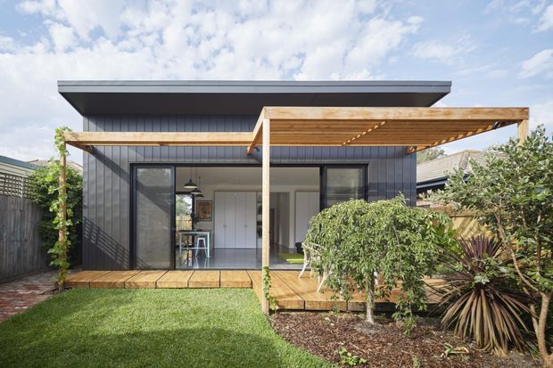 Exterior view of a compact contemporary single-storey dwelling with dark vertical cladding, a broad low-pitched roof, timber pergola and deck, extensive glazing, and a landscaped garden courtyard.