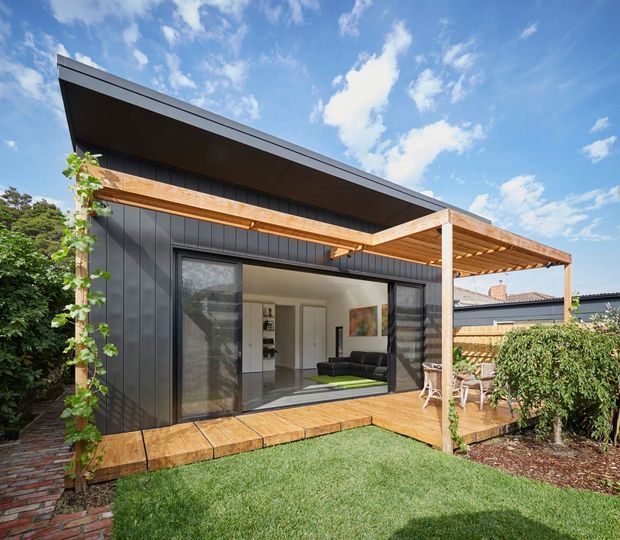 A contemporary dark-clad single-storey home extension viewed from the garden, featuring a low-pitched roof, broad timber pergola, timber decking, large sliding glass doors, and an open-plan living area visible inside. The landscaped yard includes lawn, garden beds, climbing greenery, and a brick pathway.