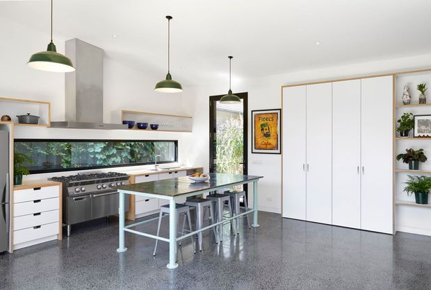 Bright contemporary kitchen with white flat-panel cabinetry, integrated open shelving, a large stainless-steel range and hood, a long horizontal garden-view window, and a central island with a dark stone benchtop and pale painted metal frame. Green pendant lights, plants, and framed artwork add restrained colour, while the polished grey concrete floor gives the room a refined industrial character.