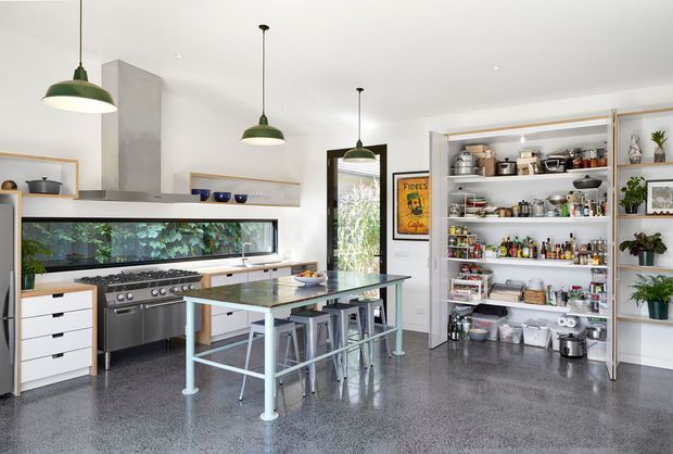 Bright, spacious contemporary kitchen with a large stainless-steel range, long horizontal window, central island with stools, polished concrete-style flooring, and an open pantry lined with shelves and supplies. White cabinetry, pale timber shelving, dark metal accents, and green pendant lights create a clean but characterful industrial-leaning interior.