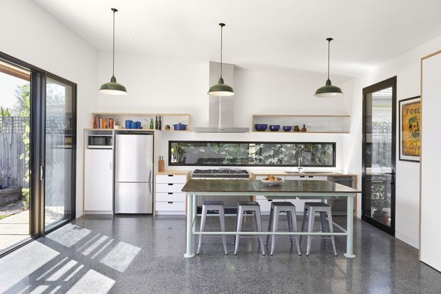 Bright contemporary kitchen with a central island and four stools, white cabinetry, stainless-steel appliances, open shelving, a long horizontal splashback window, and three green pendant lights. The room has polished grey terrazzo-style flooring, white walls, extensive glazing, and direct access to an outdoor area.
