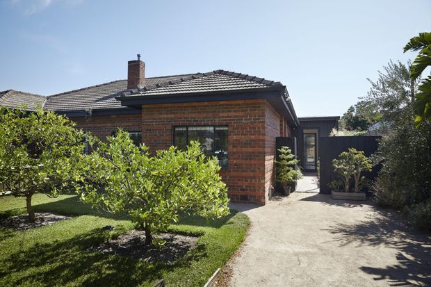Exterior view of a single-storey red-brick house with a low hipped tiled roof, prominent chimney, dark-framed windows, and a contemporary dark-clad extension or entry wing. A concrete path leads through a landscaped garden with citrus trees and clipped shrubs.