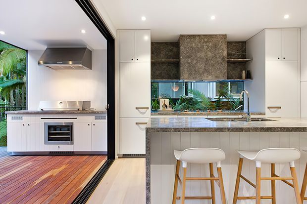 Bright contemporary kitchen with white full-height cabinetry, a large stone island with integrated seating, timber bar stools, and a textured stone feature wall and splashback. The kitchen opens directly onto an outdoor cooking or entertaining area through wide black-framed glazed doors, with greenery visible beyond.