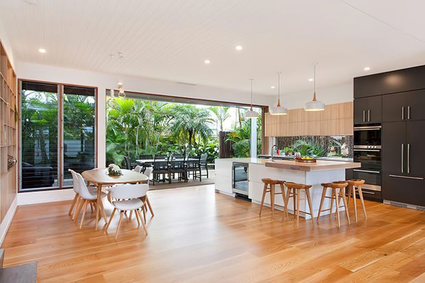 Bright, open-plan kitchen and dining area with extensive timber flooring, a large white island with timber bar stools, dark built-in kitchen cabinetry, light timber cabinetry, pendant lights, and floor-to-ceiling glazing opening to a lush outdoor area. A dining table and chairs sit beside the kitchen, creating a spacious indoor-outdoor connection.