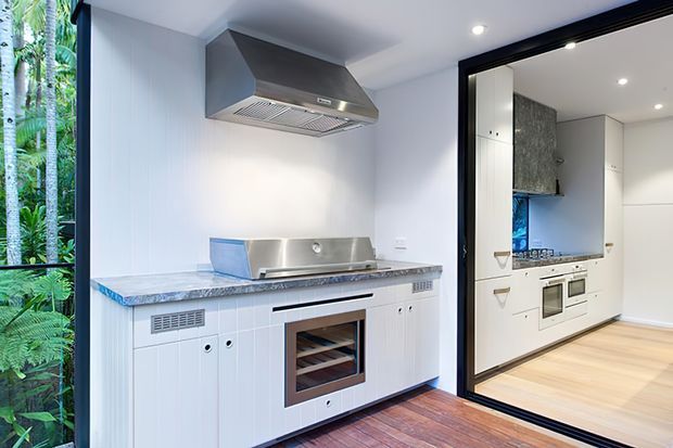 A compact, bright kitchen with white fixed cabinetry, a stainless-steel rangehood and cooktop, and a stone-look benchtop. The foreground includes a built-in oven and wine storage, while a second kitchen zone is visible through a black-framed opening. Full-height glazing on the left overlooks lush greenery, and timber flooring runs through the space.
