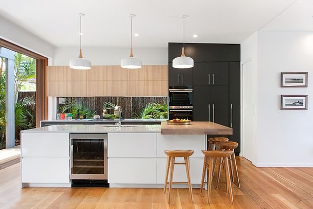 Bright contemporary kitchen with a large white island, integrated wine fridge, timber breakfast bar, light timber overhead cabinetry, dark full-height pantry cabinetry, stainless-steel appliances, and three white pendant lights. Timber flooring and a large glazed opening connect the kitchen to lush outdoor greenery.