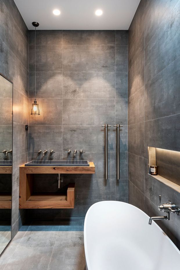 A compact contemporary bathroom featuring large-format dark grey concrete-look tiles on the walls and floor, a freestanding white bathtub, wall-mounted brushed metal tapware, a timber vanity with an integrated basin, a recessed illuminated shelf, and a suspended industrial-style pendant light.