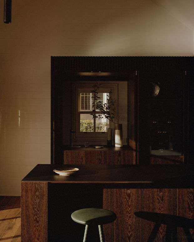 A dark, moody kitchen interior with a black stone benchtop and richly grained dark timber cabinetry. A compact island or peninsula with a rounded stool occupies the foreground, while the rear wall includes a timber-framed window, integrated joinery, open shelving, and a softly lit work area. Warm ambient lighting emphasizes the deep brown and black palette.