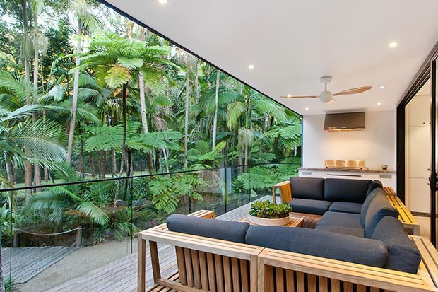 A bright, contemporary open-plan living area with a large timber-framed sectional sofa and coffee table positioned beside full-height glass balustrading and expansive glazing. Dense tropical greenery fills the view beyond the deck, while a compact kitchen or bar area with dark cabinetry and a wall-mounted television is visible at the rear. A ceiling fan, recessed lighting, timber decking, and a neutral palette create a relaxed indoor-outdoor connection.
