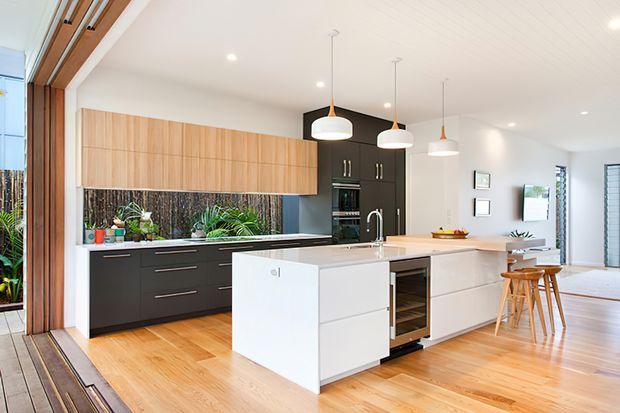 Bright contemporary open-plan kitchen with a large white island, dark lower cabinetry, warm timber overhead cabinetry, integrated appliances, timber flooring, pendant lighting, and indoor planting. The space opens toward glazed exterior doors and a dining area.