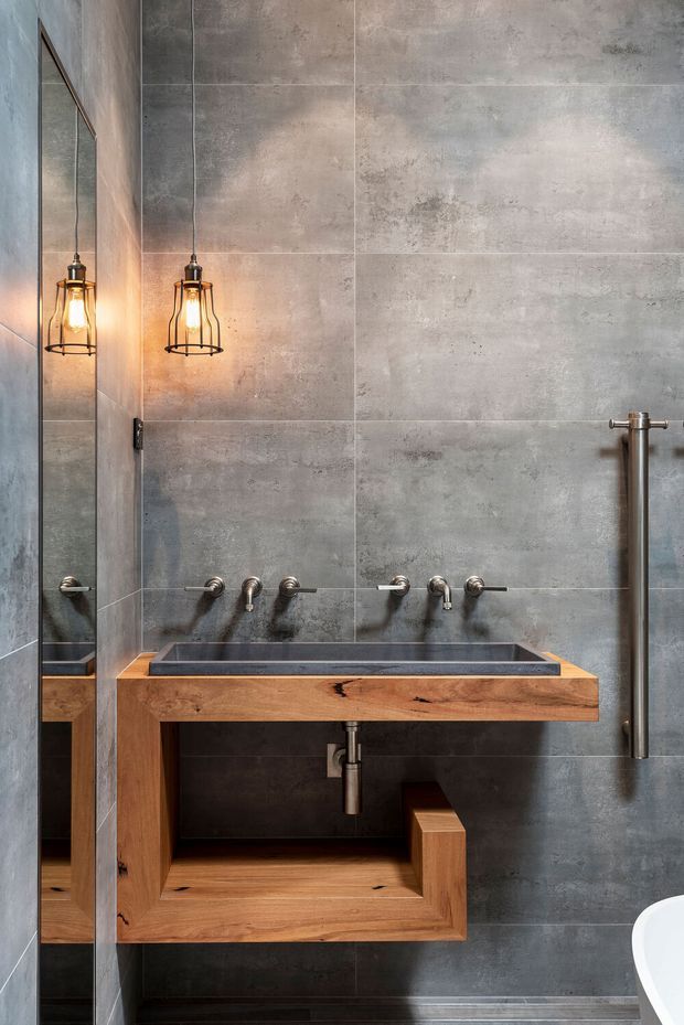 Close-up view of a contemporary industrial bathroom vanity. A wall-mounted timber vanity with an integrated dark rectangular basin sits against large grey concrete-look tiled walls. Exposed plumbing and brushed metal tapware reinforce the industrial character, while a tall mirror and warm cage pendant lights add contrast. Part of a freestanding white bath is visible at the lower right.
