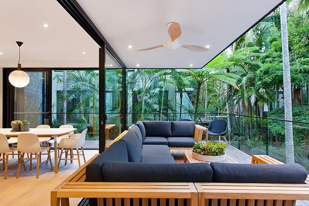 Open-plan contemporary living and dining area with a large dark sectional sofa, timber-framed outdoor-style furniture, a pale timber dining table and chairs, and full-height black-framed glazing. The space opens onto a covered terrace or balcony surrounded by dense tropical greenery, with a ceiling fan and glass balustrade visible. The interior is bright, relaxed, and strongly connected to the garden.
