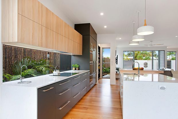 A bright contemporary kitchen with a long dark-grey island and white stone benchtops, extensive light timber overhead cabinetry, integrated dark cabinetry, and a reflective dark splashback. Warm timber flooring leads toward an open outdoor area, while pendant lights, recessed lighting, and indoor planting add a clean modern finish.