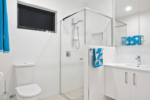 Bright, compact contemporary bathroom with a frameless glass shower enclosure, wall-mounted shower fittings, white toilet, mirrored vanity cabinet, white benchtop and chrome tapware. A small high window and blue accents add contrast to the predominantly white space.