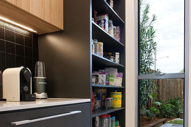 A compact, contemporary kitchen or kitchenette detail featuring dark cabinetry, a tall open pantry stocked with food items, light timber overhead cabinetry, a pale stone benchtop, and a black tiled splashback. A large window brings in natural light and overlooks a small planted courtyard.