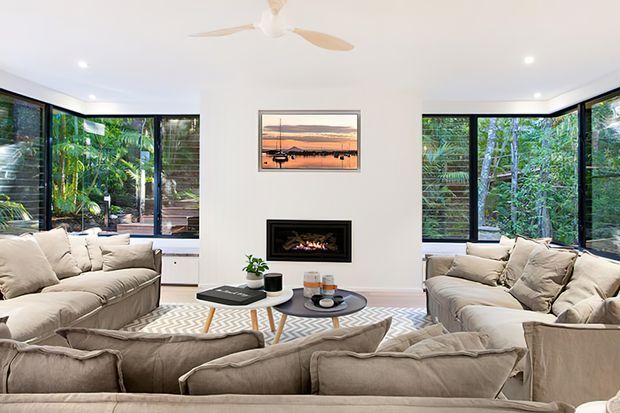 Bright contemporary living room with large corner windows overlooking lush greenery, two large neutral upholstered sofas, a geometric patterned rug, central coffee table, recessed fireplace, ceiling fan, and coastal artwork.