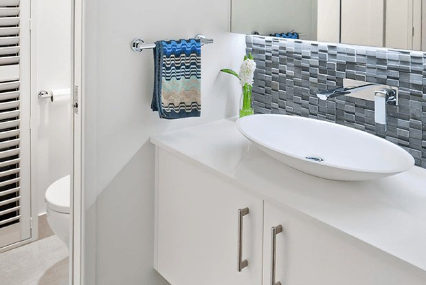 Bright contemporary bathroom vanity with a white floating-style cabinet, white vessel basin, chrome tapware, and a grey-and-white mosaic tile splashback. A mirrored cabinet reflects the basin area, while a separate toilet is partially visible through the doorway.