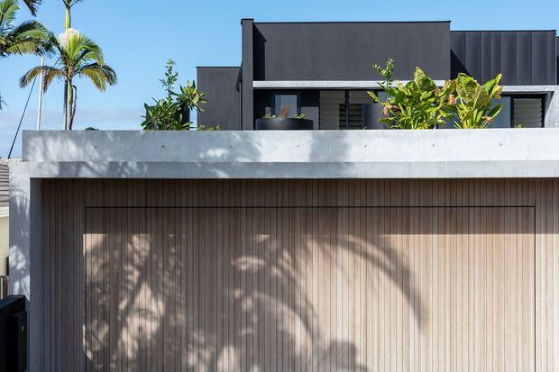 Front exterior view of a contemporary multi-level residence with a dark rendered upper facade, projecting flat roof forms, a broad pale concrete parapet, and a timber-look vertically slatted garage door. Tropical planting and palm trees frame the property, with strong sunlight creating patterned shadows across the facade.