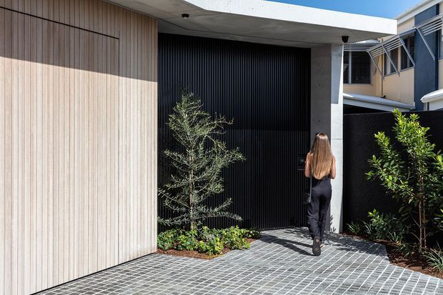 Contemporary residential entrance featuring a pale vertical timber-clad facade, a large black ribbed garage or entry door, and a broad flat concrete canopy supported by a concrete column. The approach is paved with small grey rectangular pavers and softened with planted garden beds. A neighbouring multi-level building is visible in the background.