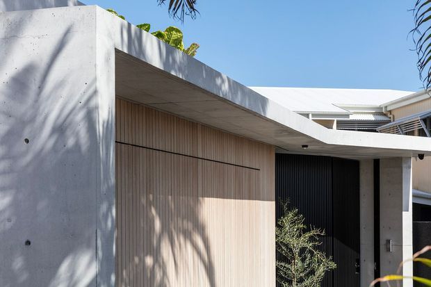 Contemporary residential facade with a minimalist flat-roofed concrete form, pale timber-look vertical garage or entry cladding, dark vertical screening, and a sheltered recessed entrance. Strong sunlight and tropical planting frame the exterior.