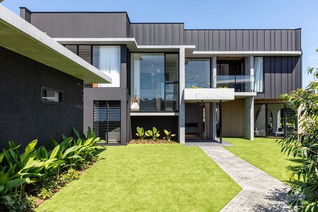 Exterior view of a contemporary multi-level residence with dark vertical cladding, smooth rendered concrete forms, extensive glazing, projecting balconies and strong horizontal roof and slab lines. A paved path crosses a manicured lawn toward the recessed entry, with tropical-style planting along the facade.