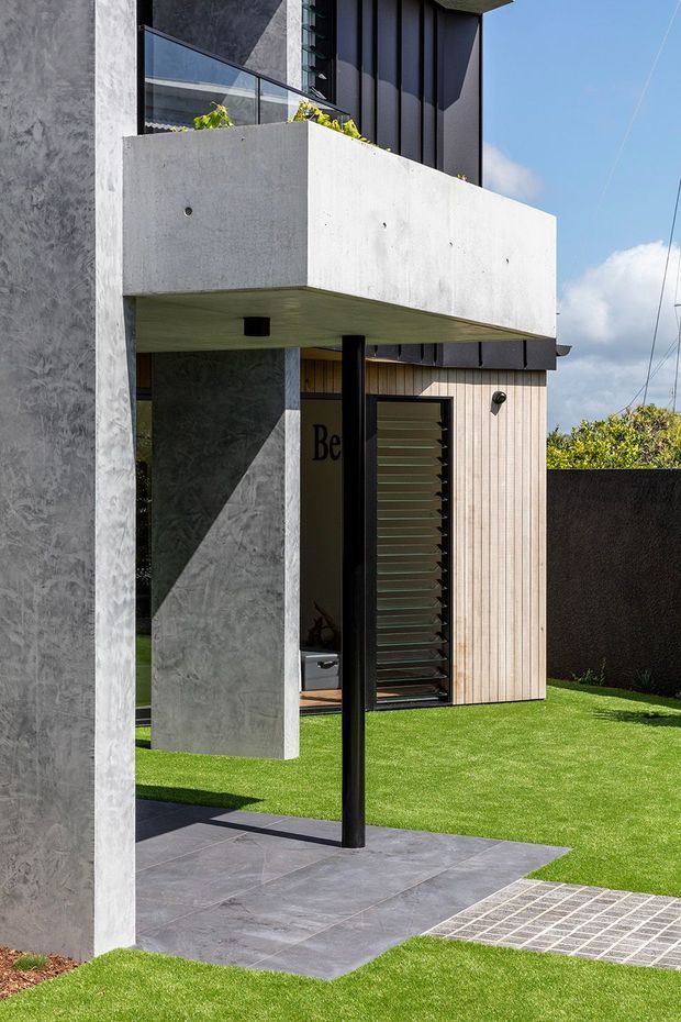 A contemporary multi-level residential facade with a projecting raw concrete balcony or canopy supported by a slender black post. The exterior combines textured grey rendered walls, dark vertical cladding, pale vertical timber-look cladding, black-framed glazing and a louvred front door. Dark large-format paving, a grated drainage strip and manicured lawn define the entry courtyard, with a small planted balcony above.