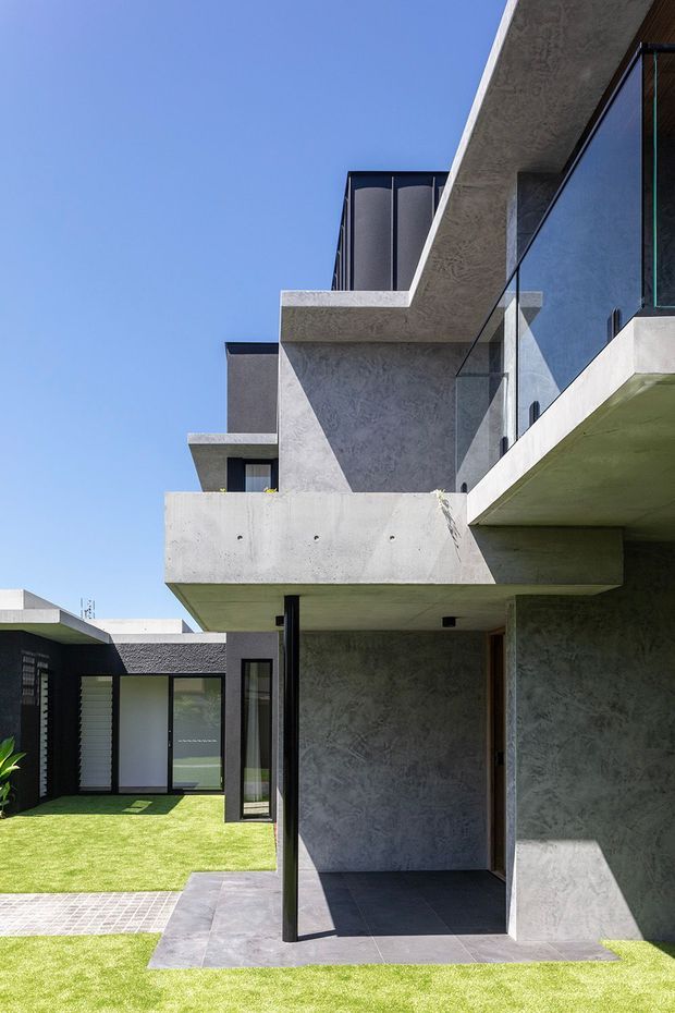 Contemporary multi-level residential exterior with strong geometric forms, cantilevered concrete balconies, dark textured rendered walls, extensive floor-to-ceiling glazing, and slim black-framed glass balustrades. The view looks across a manicured lawn and paved courtyard toward the shaded ground-level entry and adjacent single-storey wing under a clear blue sky.