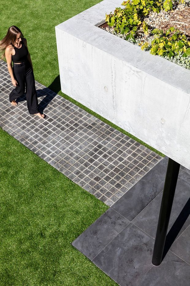 Elevated view of a contemporary landscaped outdoor area featuring a gridded dark stone or concrete paver walkway across a lawn, a large pale concrete raised planter with dense planting, and dark stone paving beneath a black structural post. A person is present for scale.