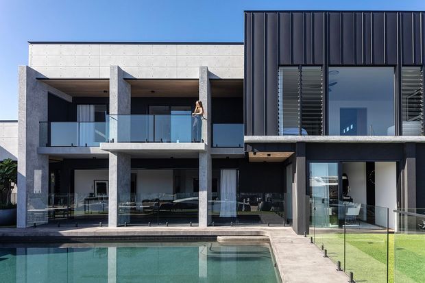 Contemporary multi-storey residential facade with strong geometric massing, raw concrete columns and balconies, dark vertical metal cladding, expansive glazing, and frameless glass balustrades. A swimming pool and manicured lawn occupy the foreground, with interior living spaces visible through the glazing.