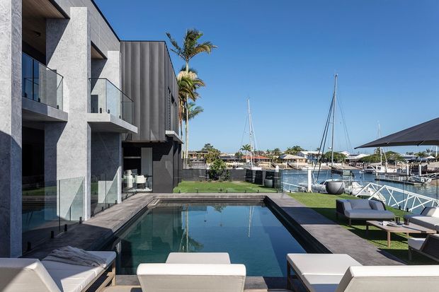 Contemporary multi-storey waterfront residence with a dark rendered and vertical-panel facade, projecting balconies with clear glass balustrades, and a long rectangular swimming pool extending through the foreground terrace. Outdoor lounge furniture surrounds the pool, while yachts and a marina are visible beyond the property, creating a bright coastal setting.