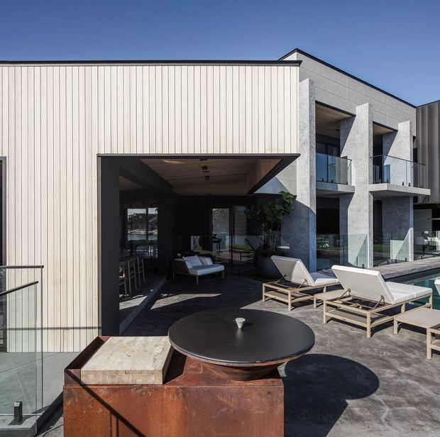 Contemporary multi-level building exterior with a rooftop terrace and outdoor lounge area. The facade combines pale vertical cladding, grey textured masonry, extensive glazing, projecting balconies, and a recessed covered outdoor space. A rusted steel outdoor kitchen or fire feature, round table, glass balustrades, and sun loungers occupy the dark paved terrace.