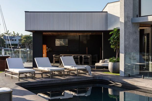 Contemporary outdoor pool terrace beside a dark, open-sided entertainment pavilion. The scene features streamlined white loungers, a dining and lounge area, timber-lined soffit, dark wall finishes, concrete architectural elements, glass balustrades, sculptural planting, and a reflective swimming pool. The setting appears bright, refined, and resort-like.